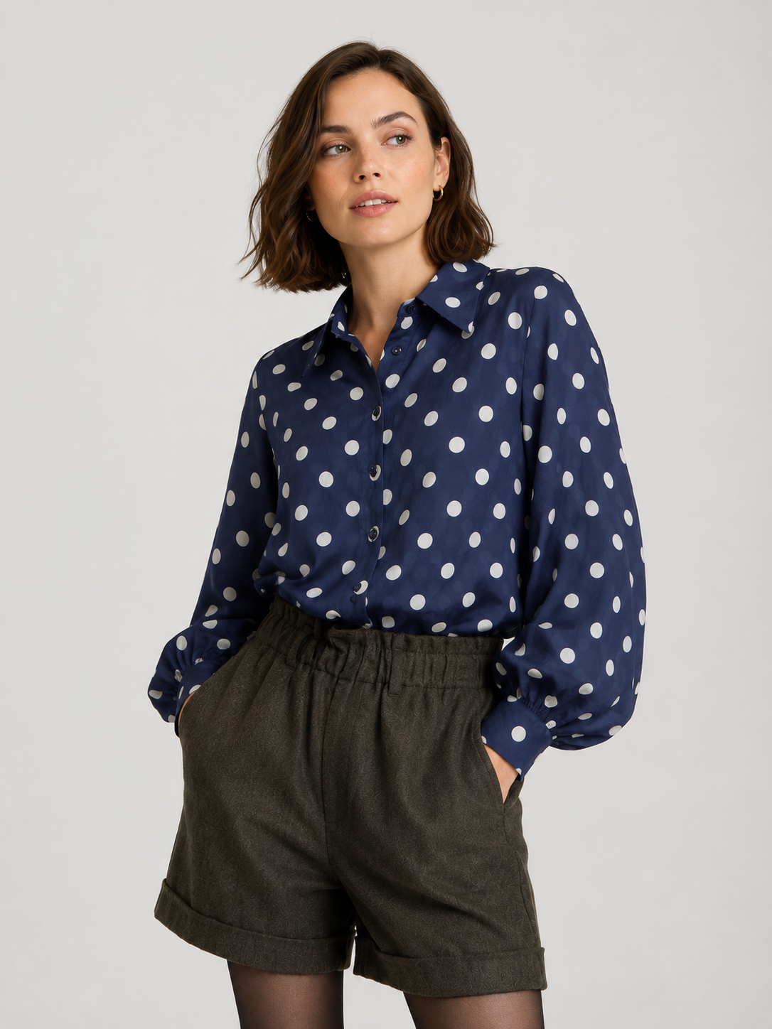 Eine Frau steht vor einem schlichten, hellen Hintergrund, die Hände in den Taschen ihrer grauen Shorts mit hoher Taille, und trägt die Oversize-Bluse „Antonia Dots Navy Ecru“ – gefertigt aus luftigem Baumwoll-Voile mit einem marineblau-weißen Polka-Dot-Muster. Hergestellt in Italien.