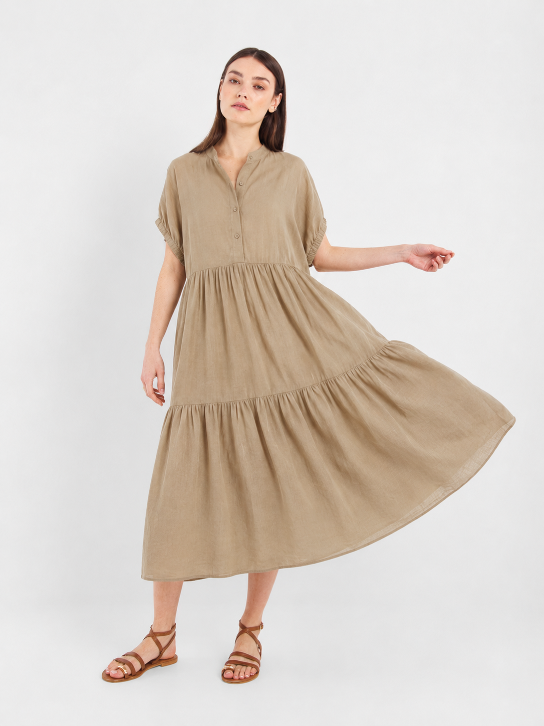Eine Frau trägt das Kleid „Cowboy Boho Sand“ – ein locker sitzendes, hellbraunes Boho-Sommerkleid mit kurzen Ärmeln, einem gestuften Rock und Knopfverzierungen am Oberteil. Das aus weicher Baumwolle gefertigte Kleid kombiniert sie mit braunen Riemchensandalen und posiert vor einem schlichten weißen Hintergrund.
