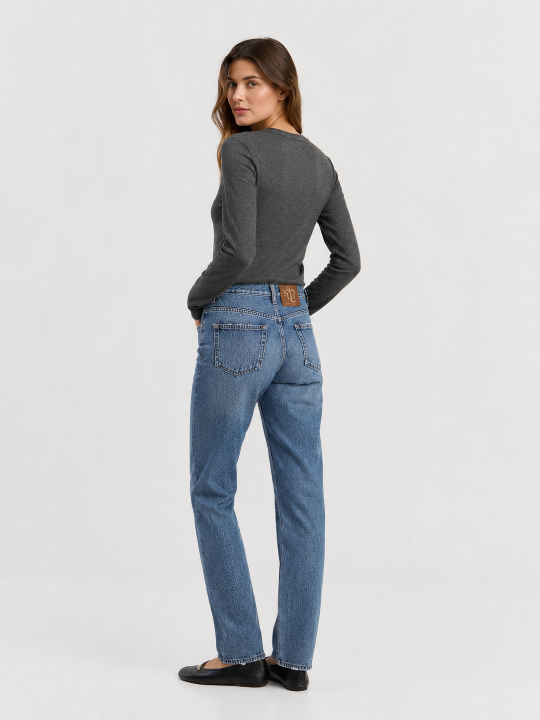 Eine Person steht mit dem Rücken zur Kamera vor einem hellen Hintergrund und trägt das dunkle Langarm-Rippshirt aus Kaschmir, blaue Jeans und schwarze Schuhe. Das Langarm-Rippshirt aus Kaschmir, das fachmännisch gefertigt und in Portugal hergestellt wurde, kommt besonders gut zur Geltung, während die Person über die Schulter in Richtung Kamera blickt.