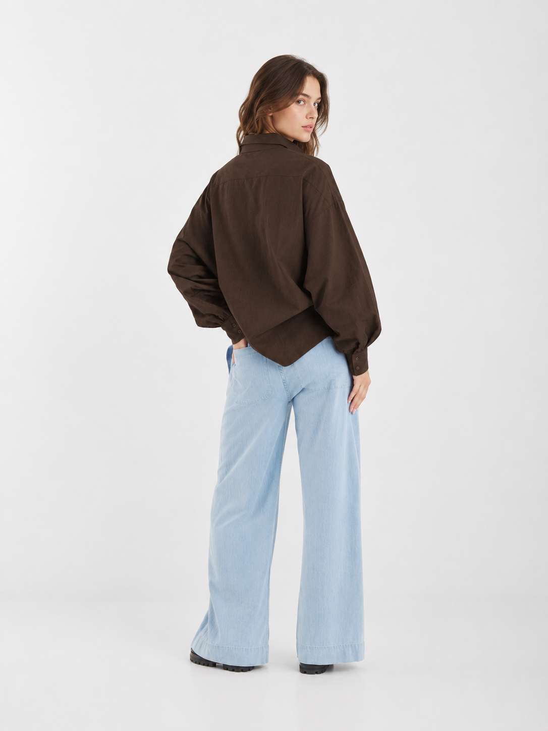 Eine Person, die eine braune Jacke über der Bluse „Antonia Slim Espresso“ mit eleganten Ballonärmeln sowie hellblaue Jeans mit weitem Bein trägt, steht mit dem Rücken zur Kamera und dreht sich leicht um, um über die Schulter zu blicken – vor einem schlichten weißen Hintergrund.