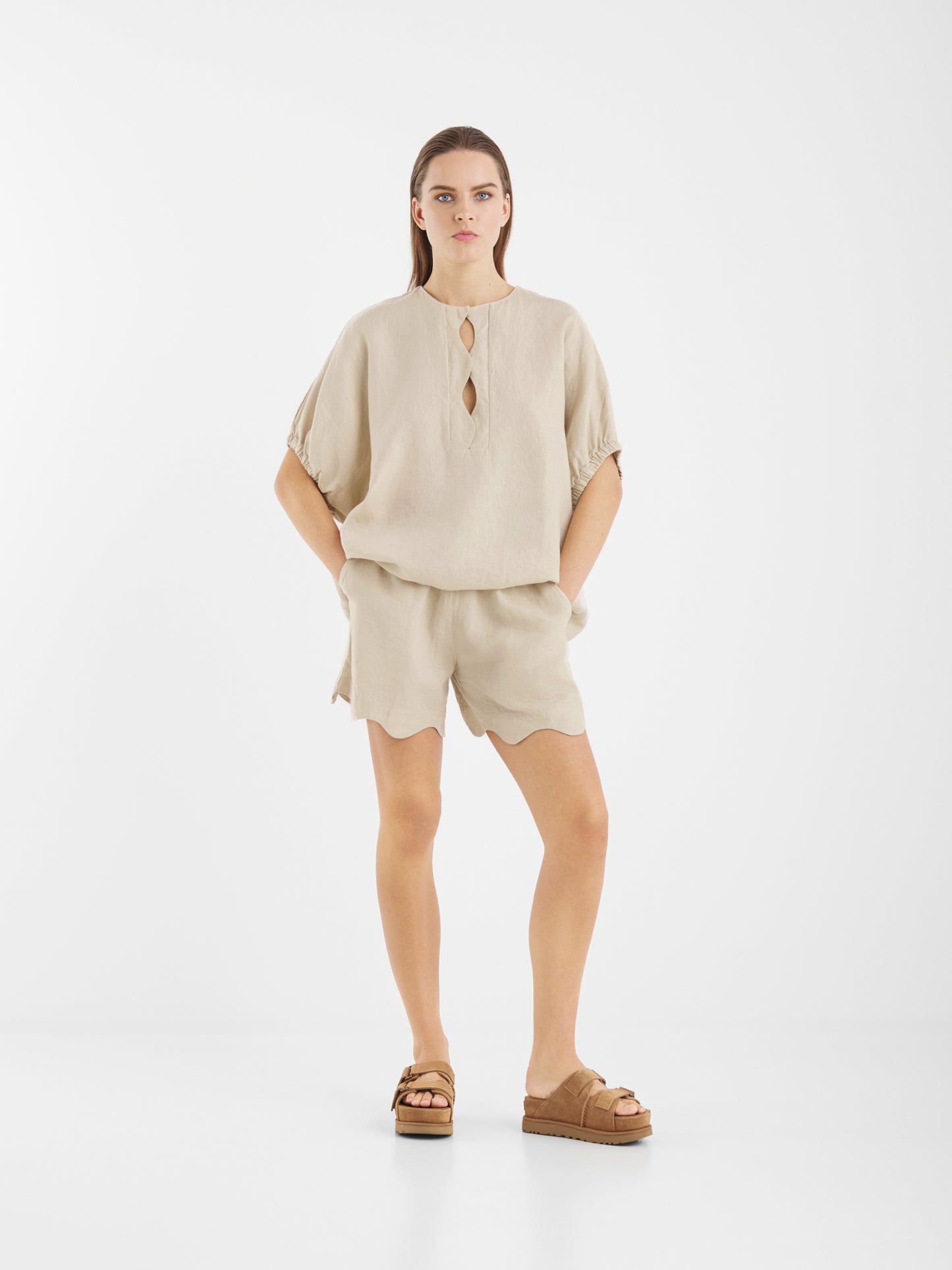Eine Frau steht vor einem weißen Hintergrund und trägt die Bluse Cowboy Wave Linen Beige mit Schlüssellochöffnung, passende Shorts, die Hände in den Taschen und braune Plateausandalen.