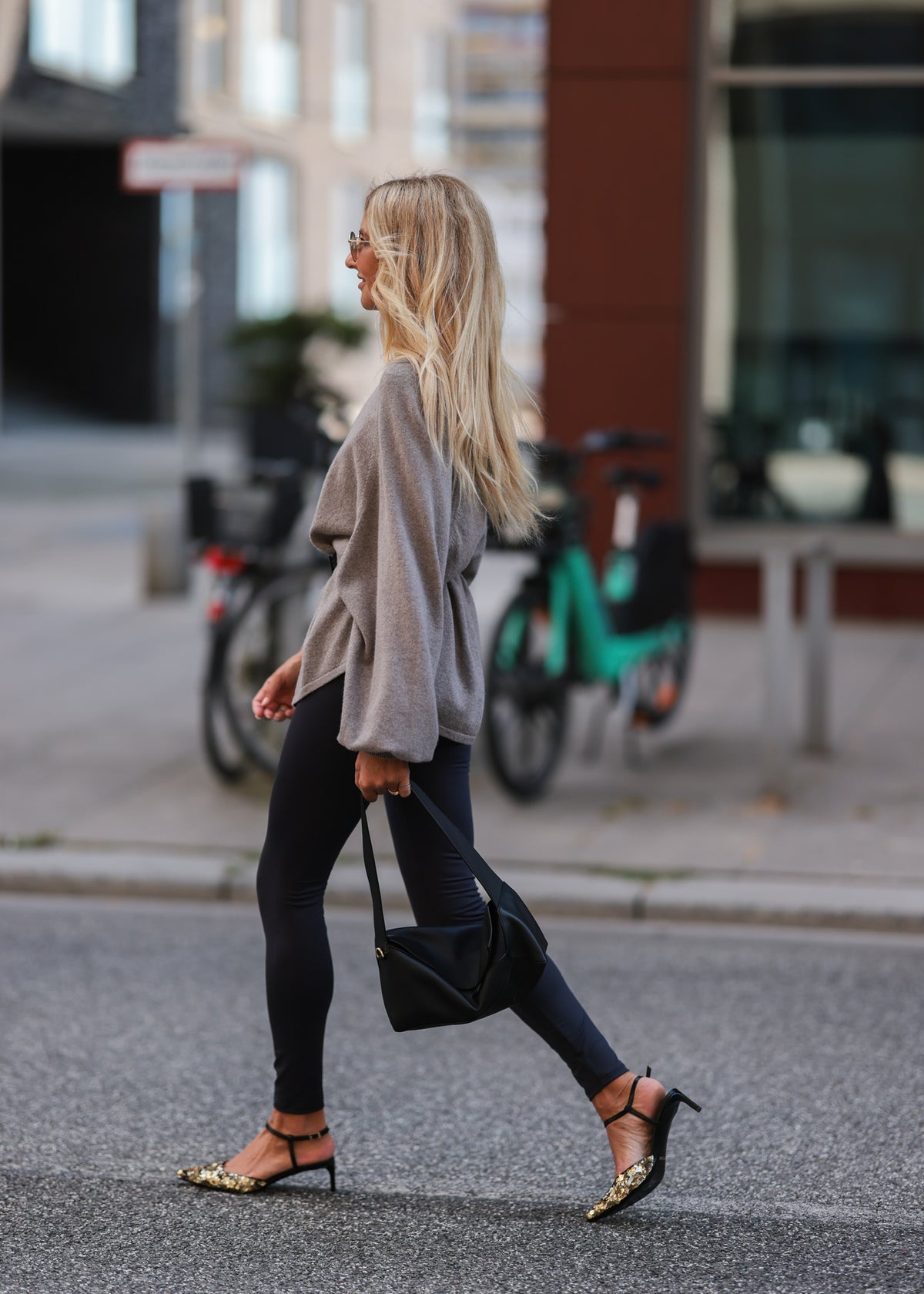 Eine Frau mit langen blonden Haaren, gekleidet in einen weiten grauen Pullover und graue Leggings aus recyceltem Polyester, geht auf einer Straße in der Stadt spazieren. Sie trägt High Heels mit Leopardenmuster und eine schwarze Tasche. Im Hintergrund sind Fahrräder und Gebäude zu sehen.