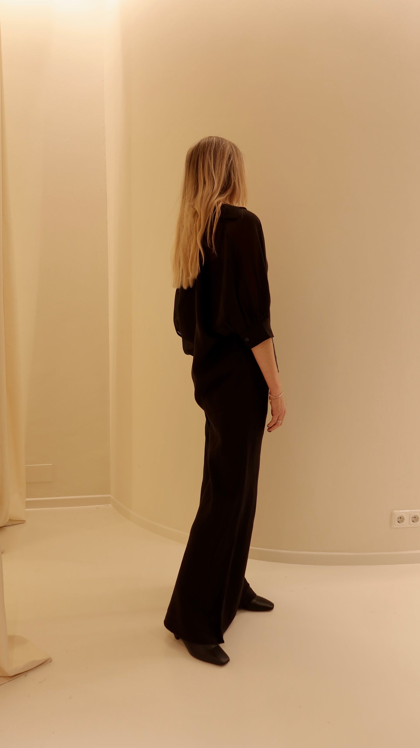 Eine Person mit langen blonden Haaren, die eine lockere Bluse Antonia Short Sleeve Black und schwarze Schuhe trägt, steht in einem minimalistischen beigen Raum und blickt von der Kamera weg leicht in die Ecke.