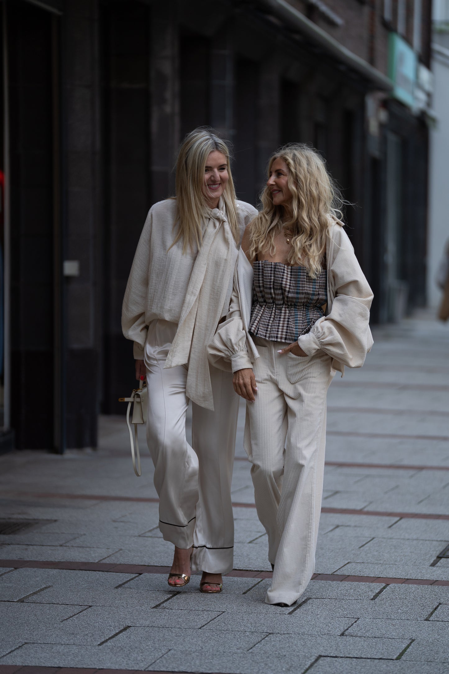 Zwei Frauen schlendern Arm in Arm die Stadtstraße entlang, beide lächeln in schicken Cremeweiß-Looks. Ihre Cordhosen 70er Jahre OFW heben sich mit lockeren Oberteilen von den dunklen Gebäuden und dem gepflasterten Bürgersteig ab.