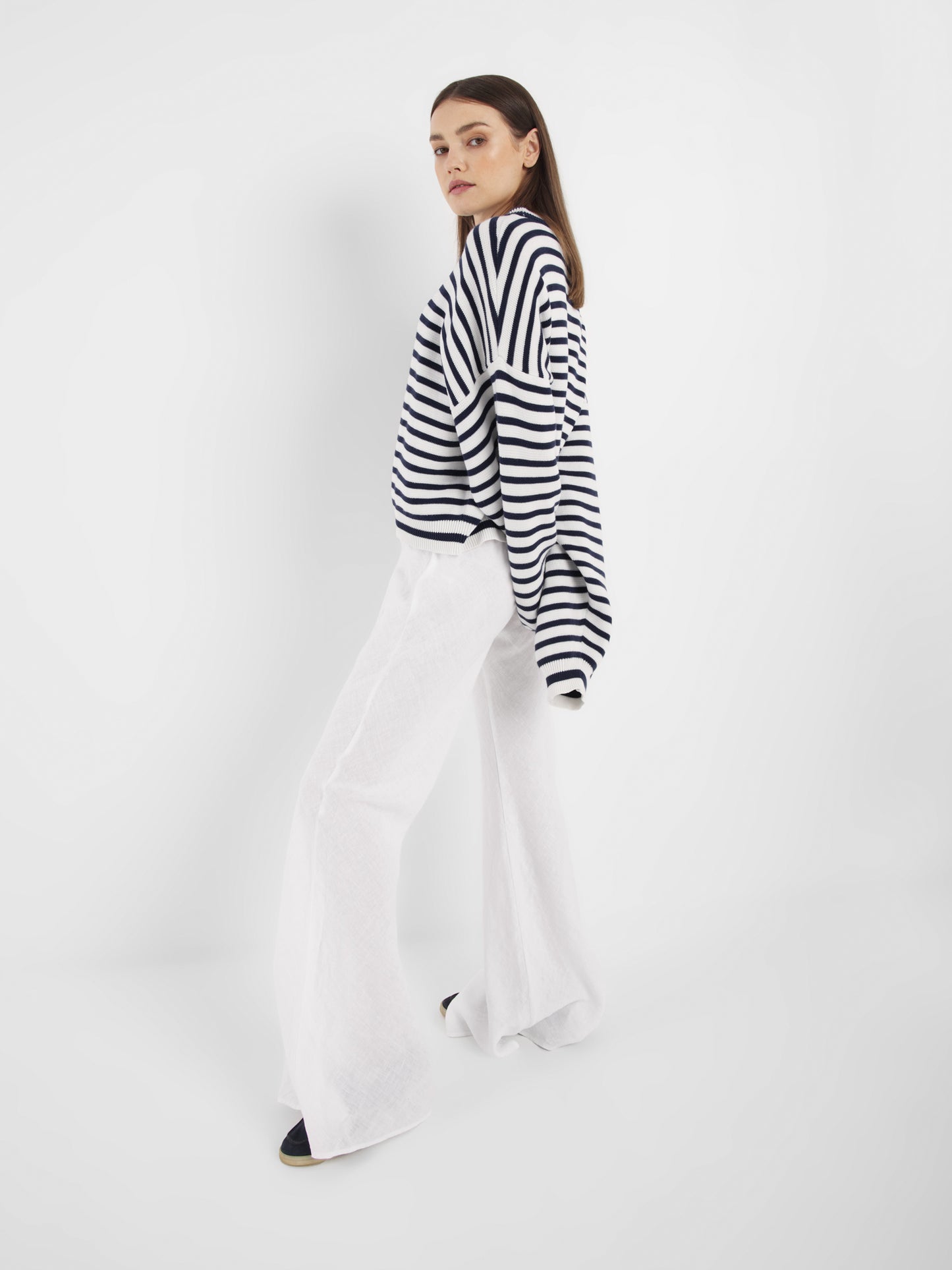 Eine Frau steht vor einem weißen Hintergrund, trägt den Pullover Sailor Navy White und eine weiße Hose mit weitem Bein und schaut mit neutralem Blick über die Schulter.