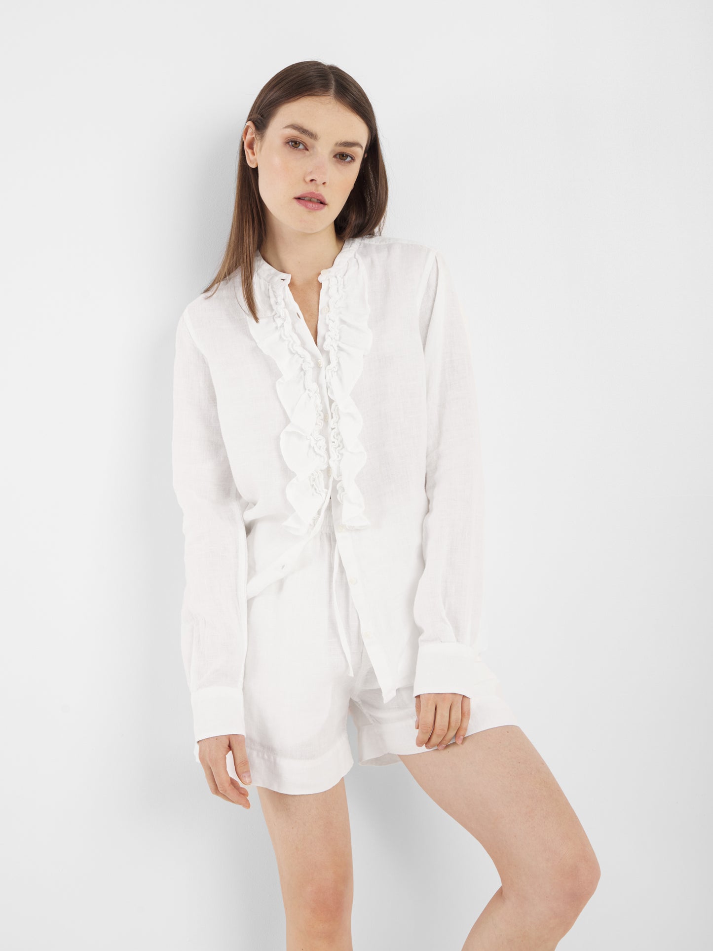 Eine Frau mit langen, glatten braunen Haaren trägt die Bluse Ruffle Linen White mit Rüschendetails und dazu passende weiße Shorts. Sie steht vor einem schlichten weißen Hintergrund und lehnt sich leicht zur Seite.