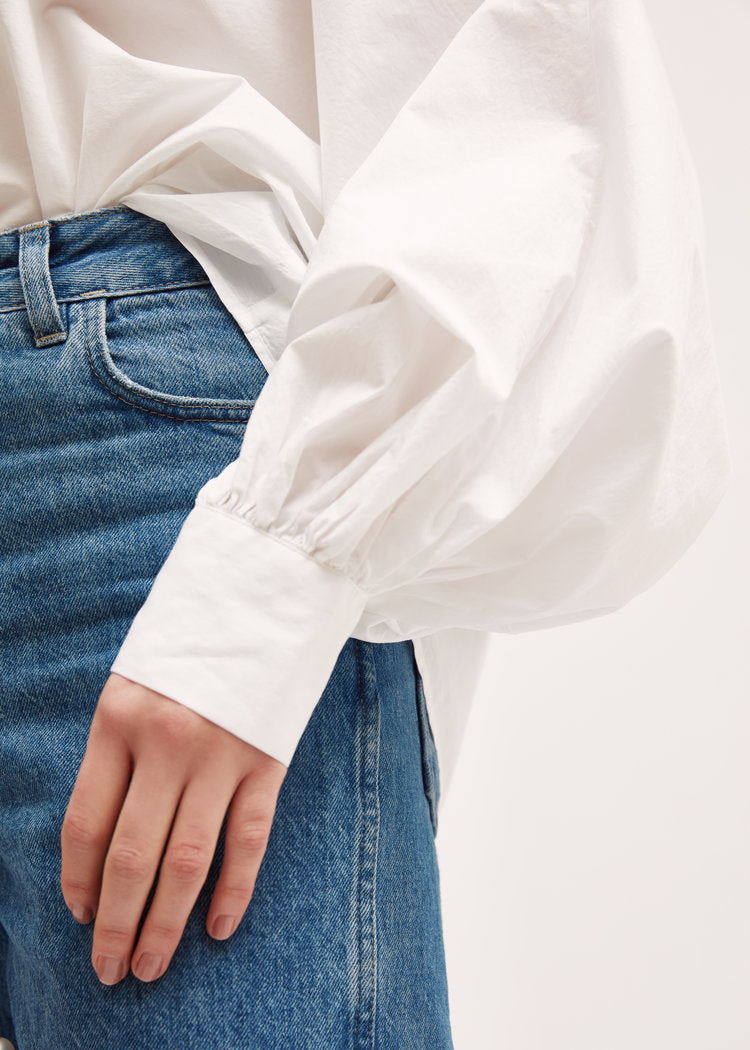 Eine Person trägt eine blaue Jeans und die Bluse Antonia White, eine übergroße Bluse mit voluminösen gerafften Ärmeln, die mit einer Hand an der Seite in der Taille gezeigt wird.
