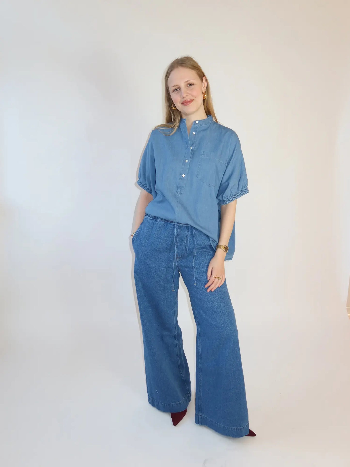 Eine Frau steht vor einem schlichten, hellen Hintergrund und trägt die Bluse Cowboy Button Denim mit chinesischem Kragen und ellenbogenlangen Ärmeln, kombiniert mit weit geschnittenen Bluejeans. Sie lächelt sanft und hat die Hände in den Taschen.