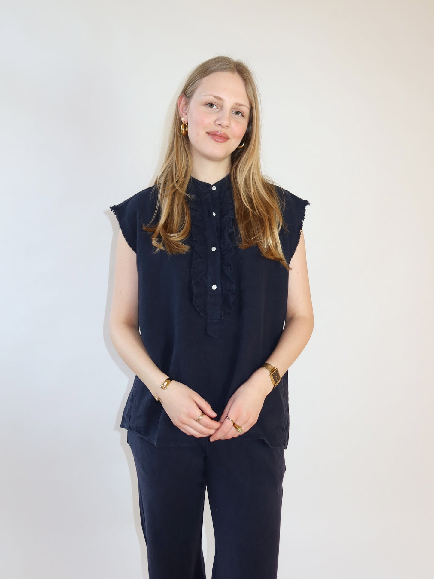 Eine junge Frau mit langem blondem Haar steht vor einem schlichten hellen Hintergrund und lächelt sanft mit gefalteten Händen. Sie trägt das Barcelona Linen Navy Set - eine ärmellose dunkelblaue Bluse mit Rüschen und passender marineblauer Hose.