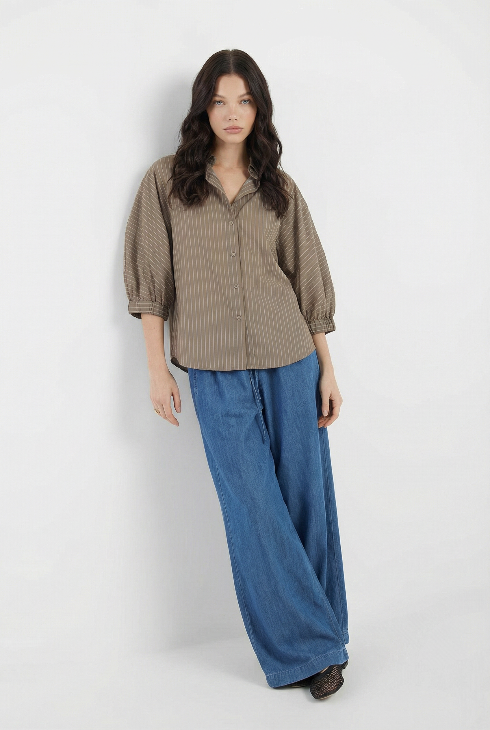 Eine Frau mit langem, dunklem, gewelltem Haar steht vor einem weißen Hintergrund. Sie trägt Anna Pants Denim mit weitem Bein und ein lockeres, braun gestreiftes Hemd mit Dreiviertelärmeln. Sie hat einen neutralen Gesichtsausdruck.