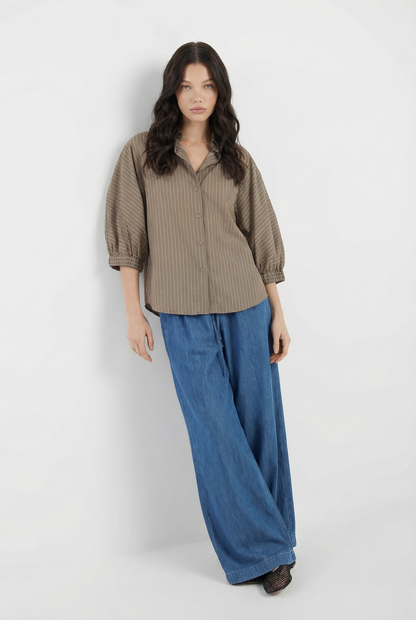 Eine Frau mit langem, dunklem, gewelltem Haar steht vor einem weißen Hintergrund. Sie trägt Anna Pants Denim mit weitem Bein und ein lockeres, braun gestreiftes Hemd mit Dreiviertelärmeln. Sie hat einen neutralen Gesichtsausdruck.