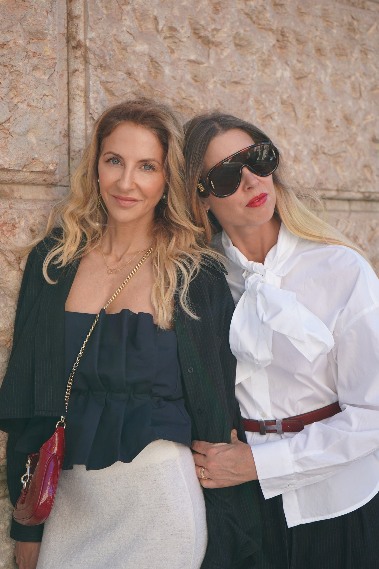 Zwei Frauen posieren an einer Steinmauer. Die eine trägt ein schickes Pleat Top Navy und eine schwarze Jacke und hat eine rote Handtasche dabei. Die andere, mit glattem blondem Haar und Sonnenbrille, trägt ein schickes Schleifentop und stützt sich auf ihre Freundin.