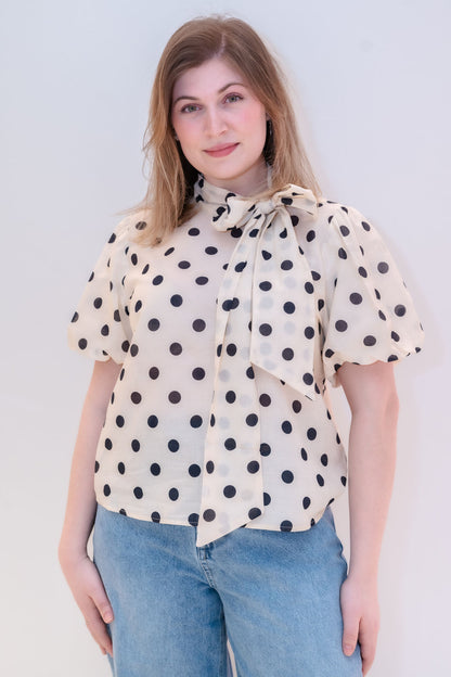 Eine Frau mit hellbraunem Haar steht vor einem hellen Hintergrund und trägt die schicke Bluse Bow Dots Ecru Black mit auffälligen schwarzen Tupfen und einer großen Schleife im Nacken, die stilvoll mit hellblauen Jeans kombiniert wird.