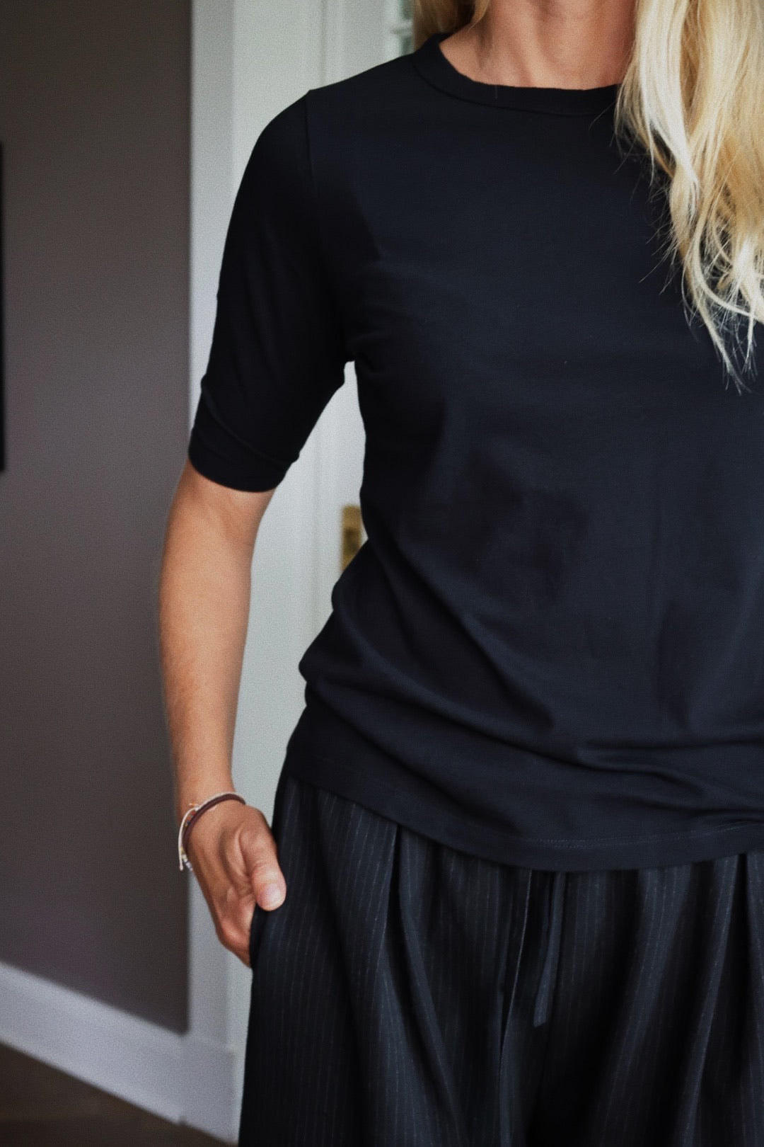 Eine Person mit langen blonden Haaren trägt ein halbarmiges schwarzes Hemd und eine dunkle Nadelstreifenhose und steht in einem Haus mit einer Hand in der Tasche. Ihr Gesicht ist nicht zu sehen.