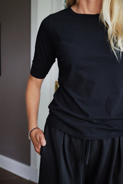 Eine Person mit langen blonden Haaren trägt ein halbarmiges schwarzes Hemd und eine dunkle Nadelstreifenhose und steht in einem Haus mit einer Hand in der Tasche. Ihr Gesicht ist nicht zu sehen.