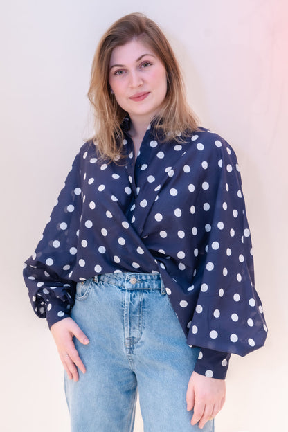 Eine Frau mit schulterlangem hellbraunem Haar lächelt sanft in die Kamera. Sie trägt die Bluse Antonia Dots Navy Ecru mit weiten Ärmeln und hellblaue Jeans und steht vor einem hellen Hintergrund.