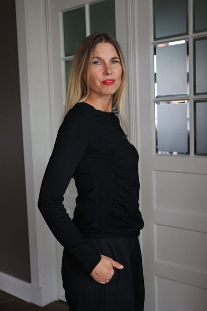 Eine Frau mit langen blonden Haaren, die ein schwarzes Hemd und eine schwarze Hose trägt, steht in einem Innenraum in der Nähe einer weißen Tür mit Glasscheiben, blickt leicht zur Seite und lächelt sanft.