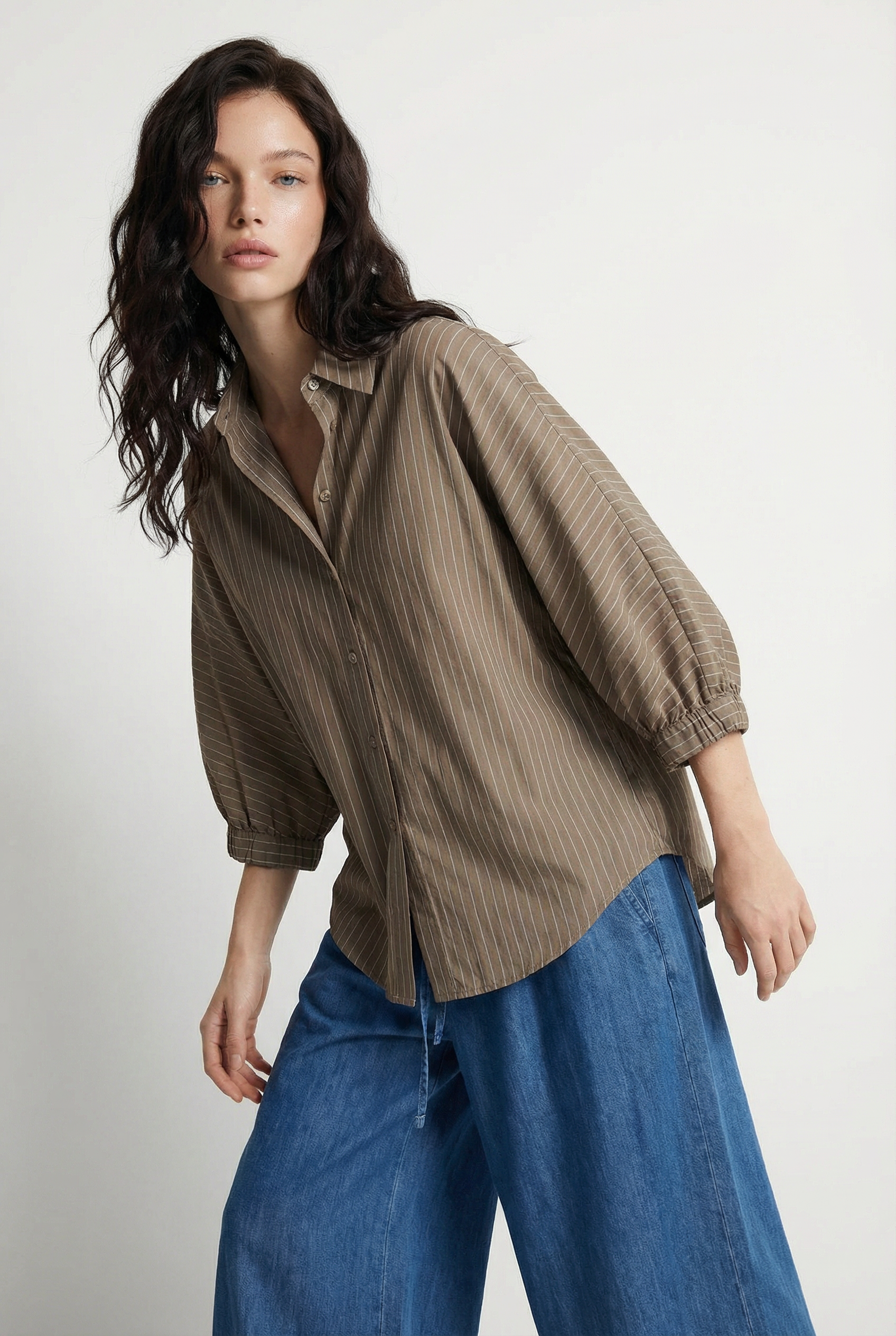 Eine Frau mit gewelltem dunklem Haar trägt eine lockere Bluse Bella Taupe Stripes mit Dreiviertelärmeln und blaue Jeans mit weitem Bein und posiert vor einem schlichten hellen Hintergrund.