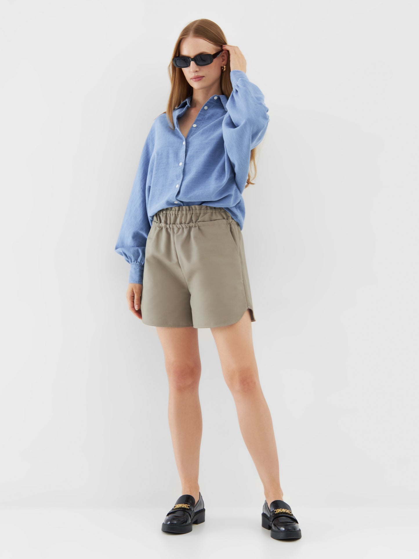 Eine Frau mit langem, glattem Haar und Sonnenbrille steht vor einem schlichten weißen Hintergrund und trägt eine Bluse Antonia Slim Denim, khakifarbene Shorts mit hoher Taille und schwarze Slipper.