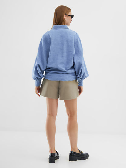 Eine Frau mit glattem, hellbraunem Haar blickt von sich weg. Sie trägt eine hellblaue Bluse Antonia Slim Denim im Oversized-Look, khakifarbene Shorts, schwarze Slipper und eine schwarze Sonnenbrille vor einem schlichten weißen Hintergrund.