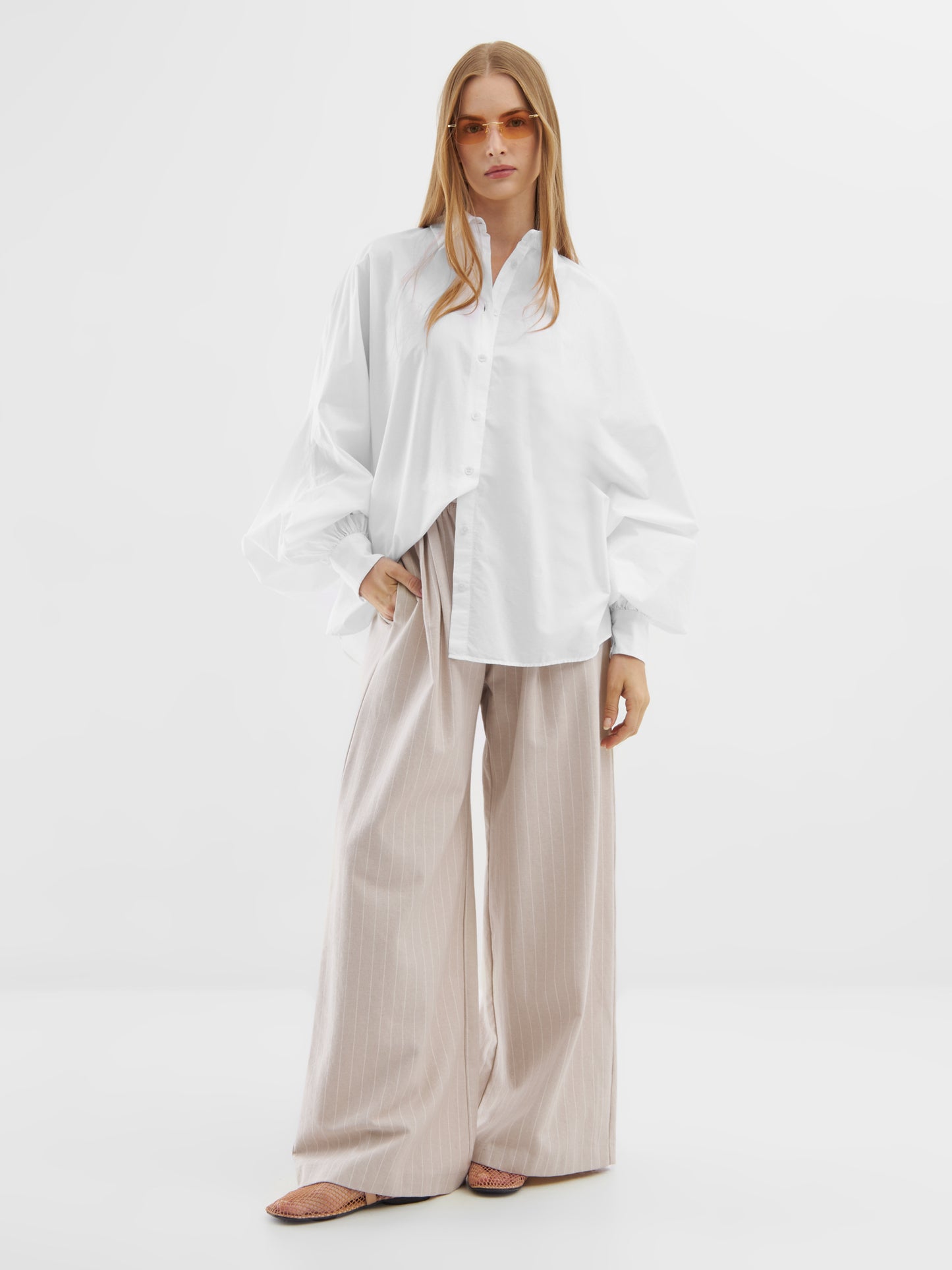 Eine Frau in der Bluse Antonia White, einer beigen Hose mit weitem Bein, einer orangefarbenen Sonnenbrille und flachen Schuhen steht selbstbewusst vor einem schlichten weißen Hintergrund.