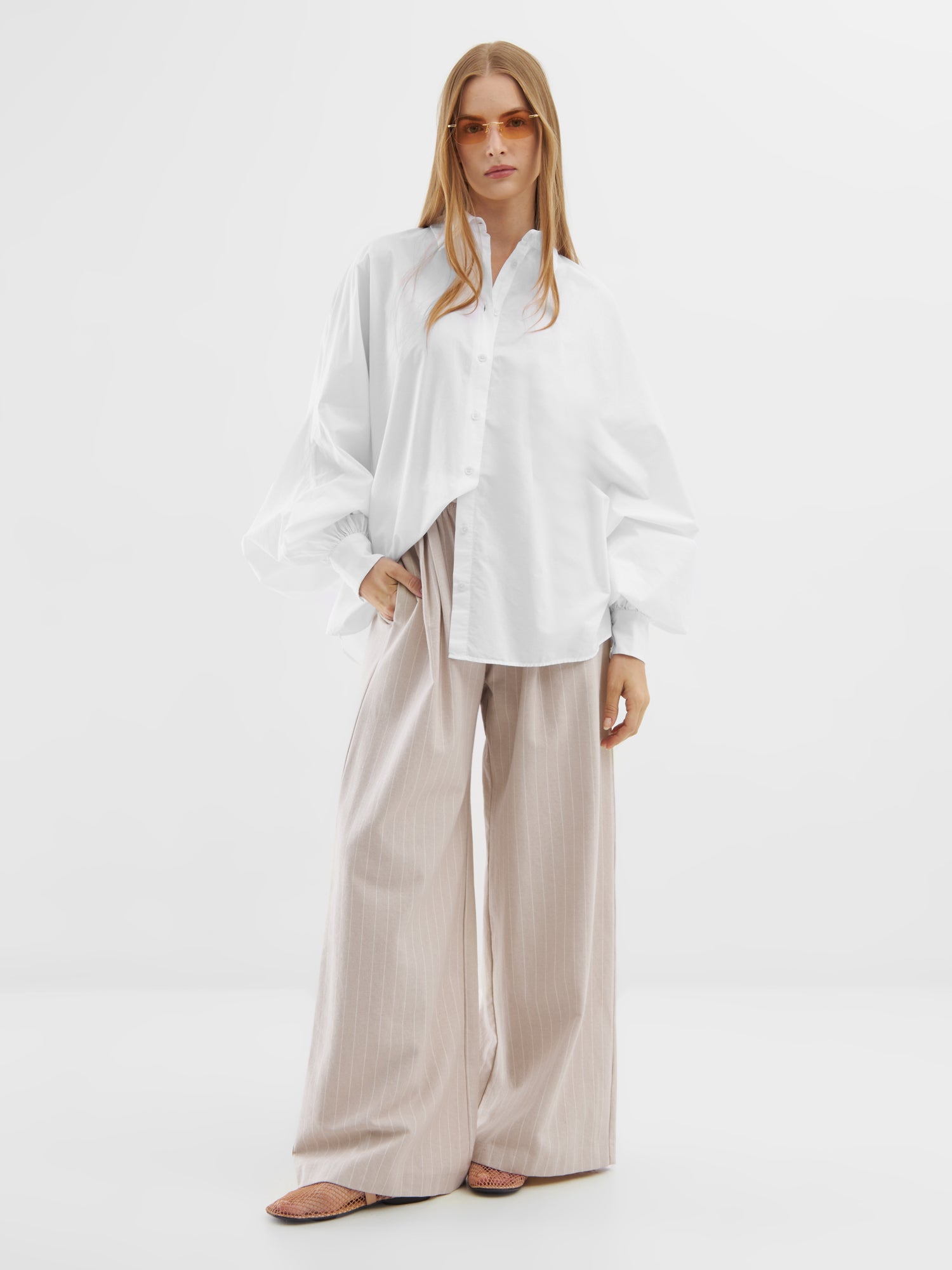 Eine Frau in der Bluse Antonia White, einer beigen Hose mit weitem Bein, einer orangefarbenen Sonnenbrille und flachen Schuhen steht selbstbewusst vor einem schlichten weißen Hintergrund.