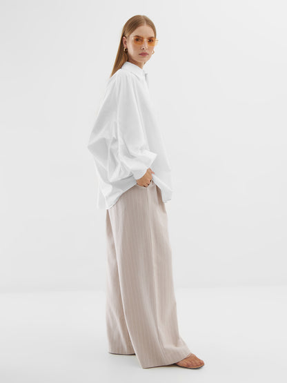 Eine Frau steht vor einem schlichten Hintergrund und trägt die Bluse Antonia White, eine beigefarbene Hose mit weitem Bein, braune offene Schuhe, goldene Ohrringe und eine leicht getönte Sonnenbrille. Ihre Hand steckt lässig in ihrer Tasche.