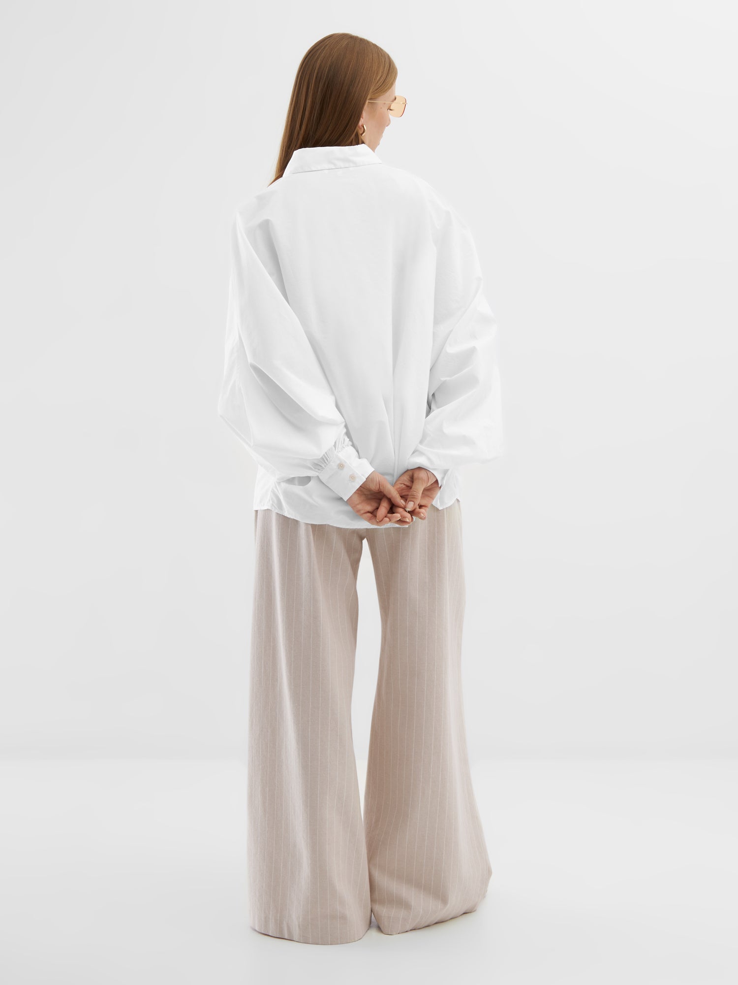 Eine Person mit langen Haaren, einer Sonnenbrille, einer übergroßen Bluse Antonia White und einer beigefarbenen Hose mit weiten Beinen steht mit hinter dem Rücken verschränkten Händen vor einem schlichten weißen Hintergrund.