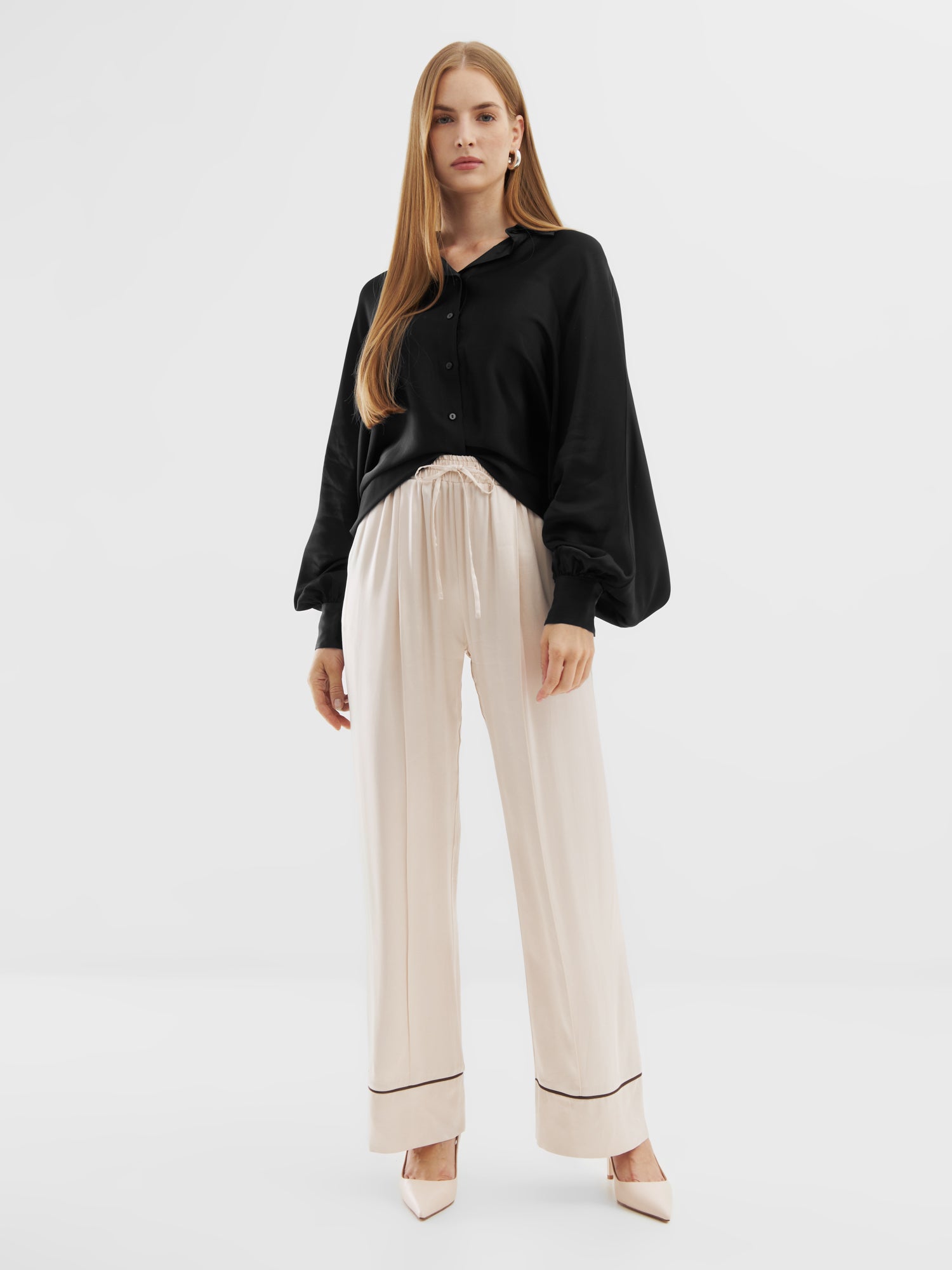 Eine Frau mit langem glattem Haar trägt eine schwarze Bluse Antonia Shiny Slim, eine hellbeige Hose mit weitem Bein und beigefarbene, spitze Absätze vor einem schlichten weißen Hintergrund.