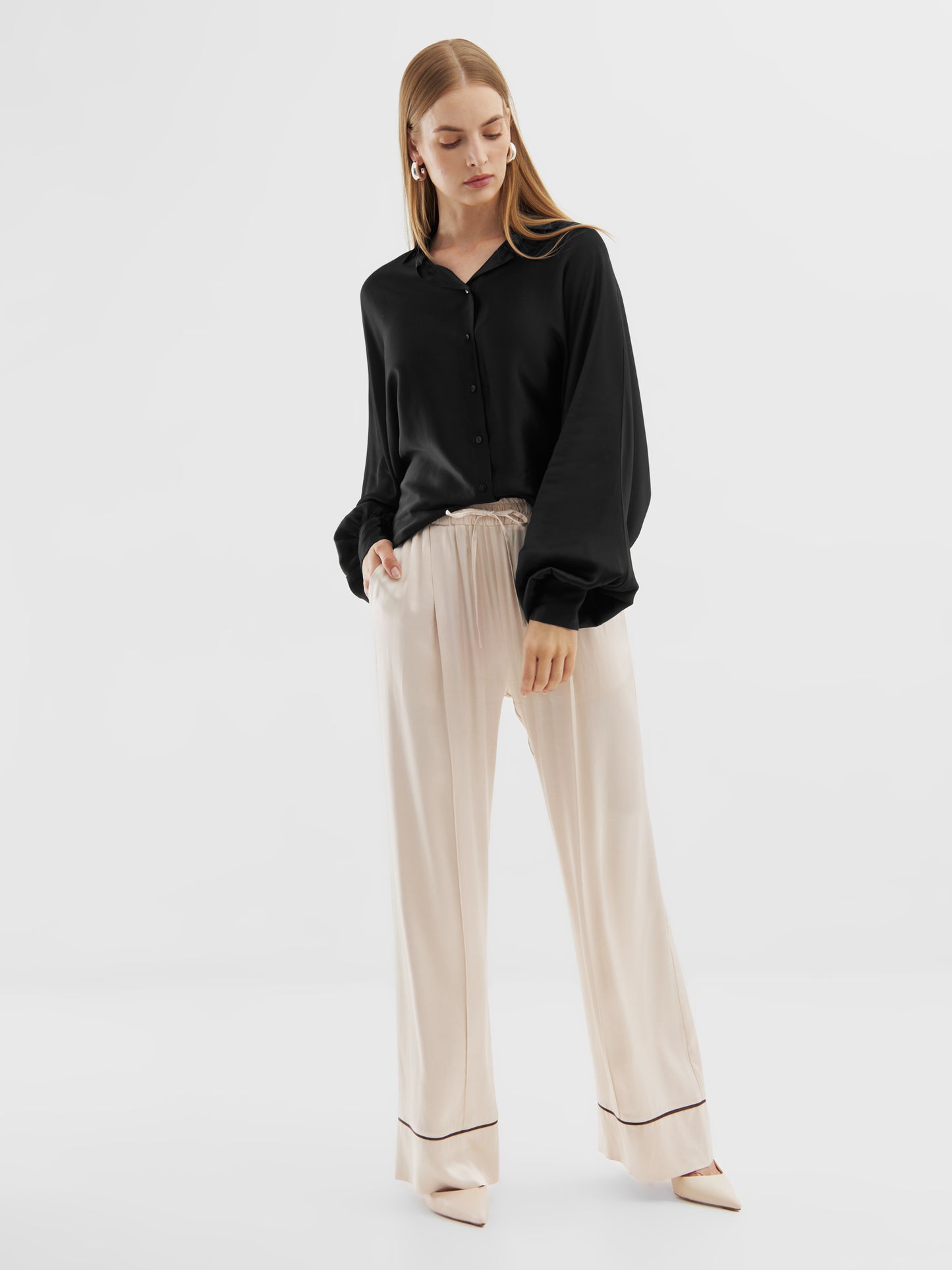 Eine Frau steht vor einem weißen Hintergrund und trägt die Bluse Antonia Shiny Slim Black mit weiten Ärmeln, eine beigefarbene Hose mit weitem Bein und dunklem Saum sowie spitz zulaufende Schuhe. Sie blickt nach unten und umarmt einen übergroßen Look mit einer Hand in ihrer Tasche.