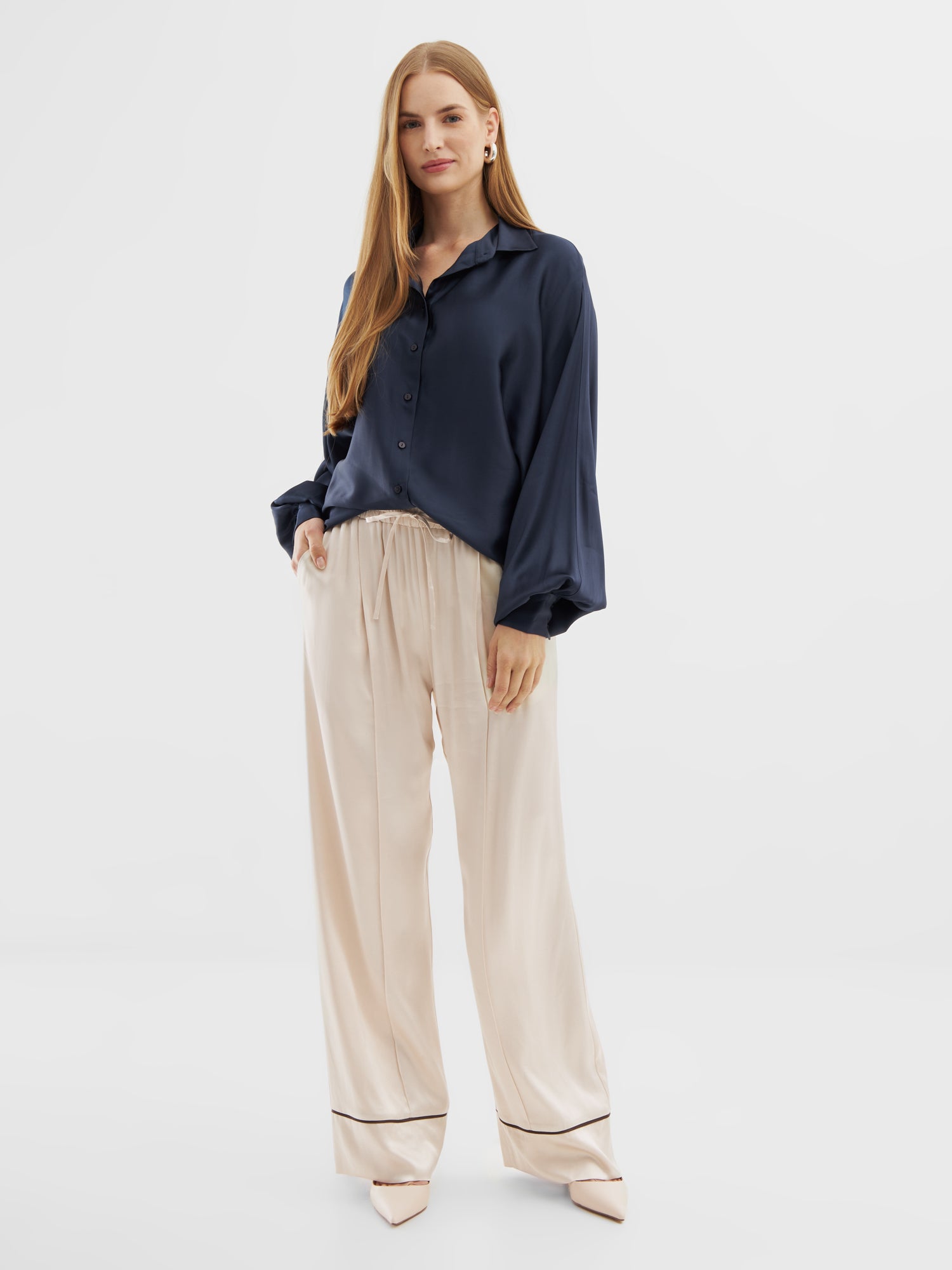 Eine Frau mit langem, glattem Haar trägt die Bluse Antonia Shiny Slim Navy und eine hellbeige Satinhose mit weitem Bein und schwarzen Paspeln am Saum, gepaart mit beigen spitzen Absätzen, vor einem schlichten weißen Hintergrund.