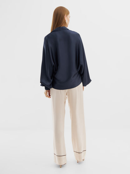 Eine Frau steht mit dem Gesicht zur Wand und trägt die Bluse Antonia Shiny Slim Navy mit langen Ärmeln und einer cremefarbenen Hose mit weitem Bein, die am Saum dunkel abgesetzt ist, vor einem schlichten weißen Hintergrund.