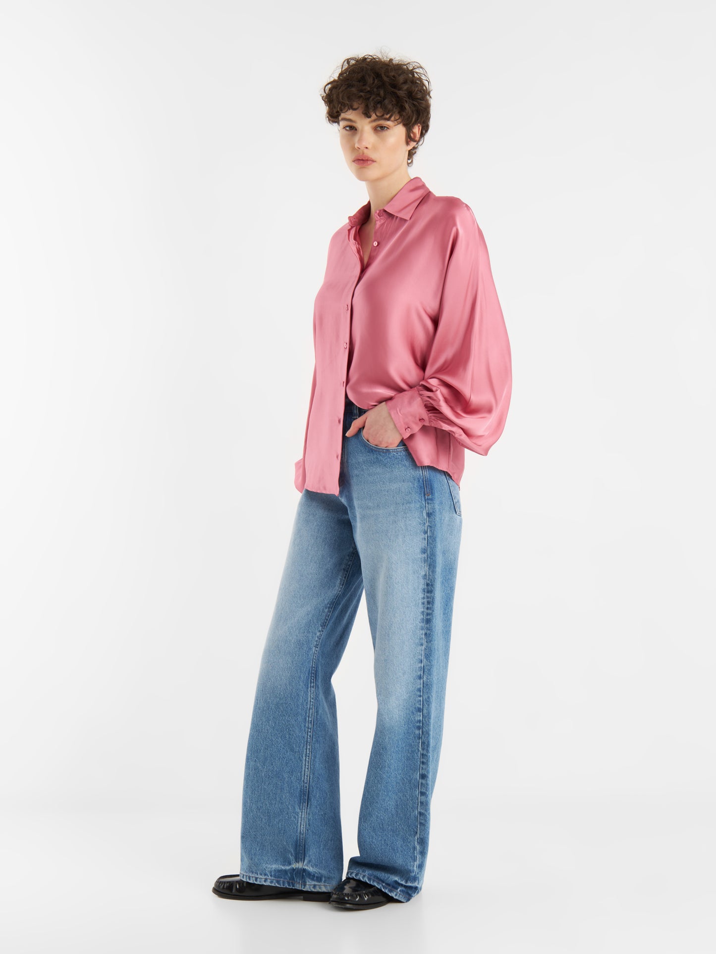 Eine Person mit kurzen lockigen Haaren trägt eine Bluse Antonia Shiny Slim Rosewood, lange Ärmel, gepaart mit hellblauen Jeans mit weitem Bein und schwarzen Schuhen, vor einem schlichten weißen Hintergrund.