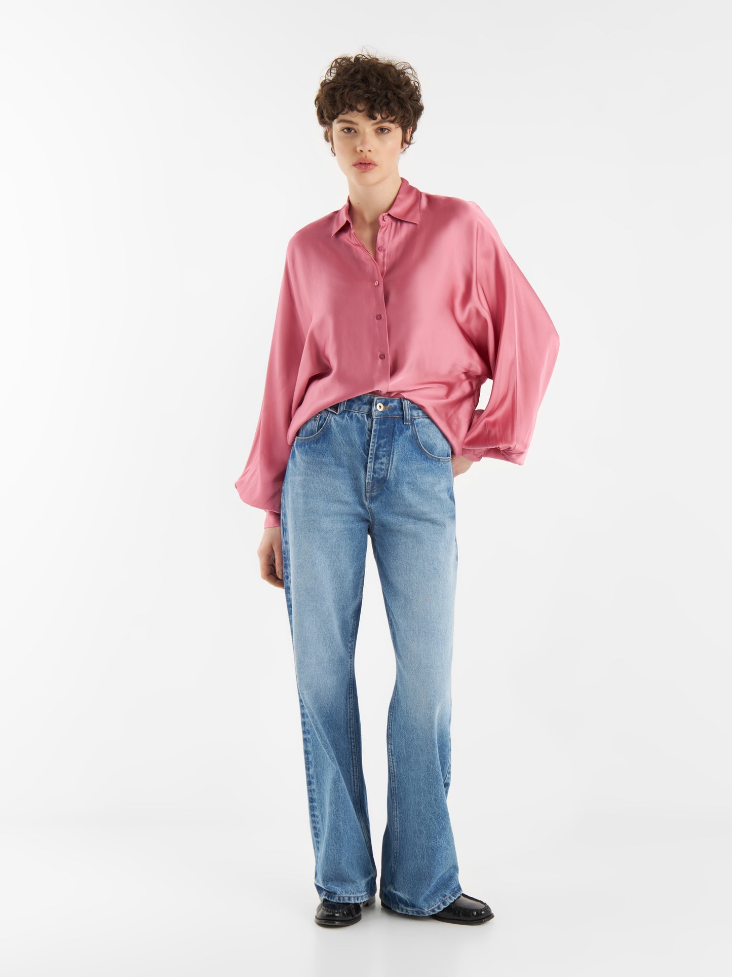 Eine Person steht vor einem weißen Hintergrund und trägt eine Bluse Antonia Shiny Slim Rosewood, hellblaue Jeans mit weitem Bein und schwarze Schuhe. Sie hat kurzes, lockiges Haar und posiert entspannt mit einer Hand an der Seite.