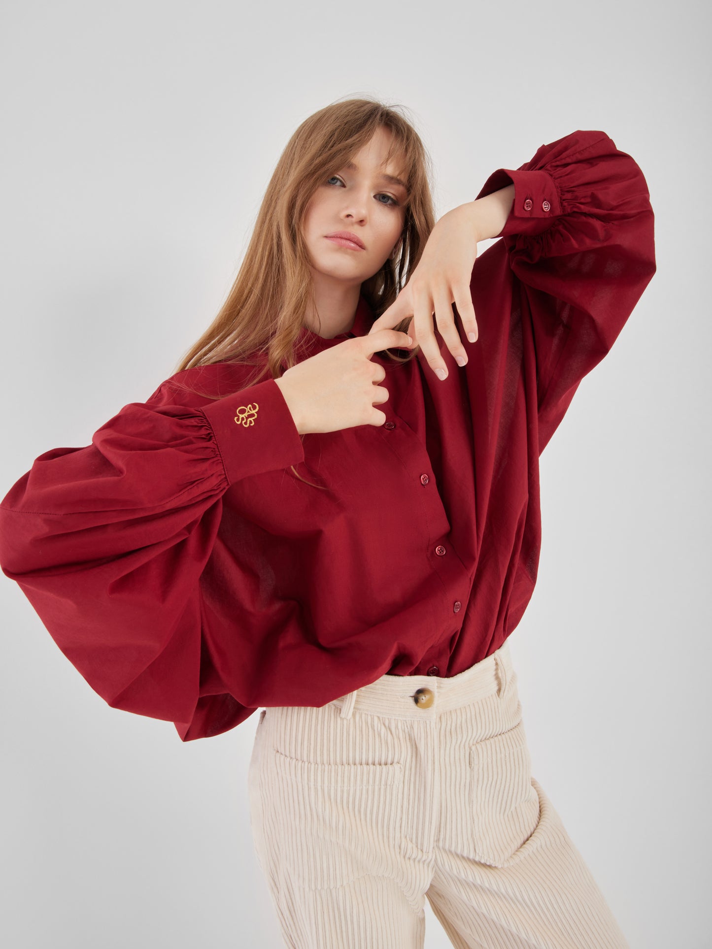 Eine Frau mit langen hellbraunen Haaren posiert in einer lockeren Bluse Antonia Burgundy mit voluminösen Ärmeln und einer cremefarbenen Hose vor einem schlichten hellgrauen Hintergrund.