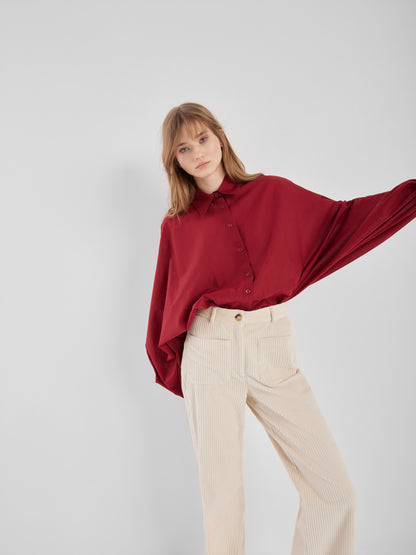 Eine Frau steht vor einem schlichten, hellen Hintergrund und trägt die locker sitzende Bluse Antonia Burgundy mit weiten Ärmeln, gepaart mit einer cremefarbenen Hose mit hoher Taille. Sie schaut mit neutralem Blick in die Kamera.