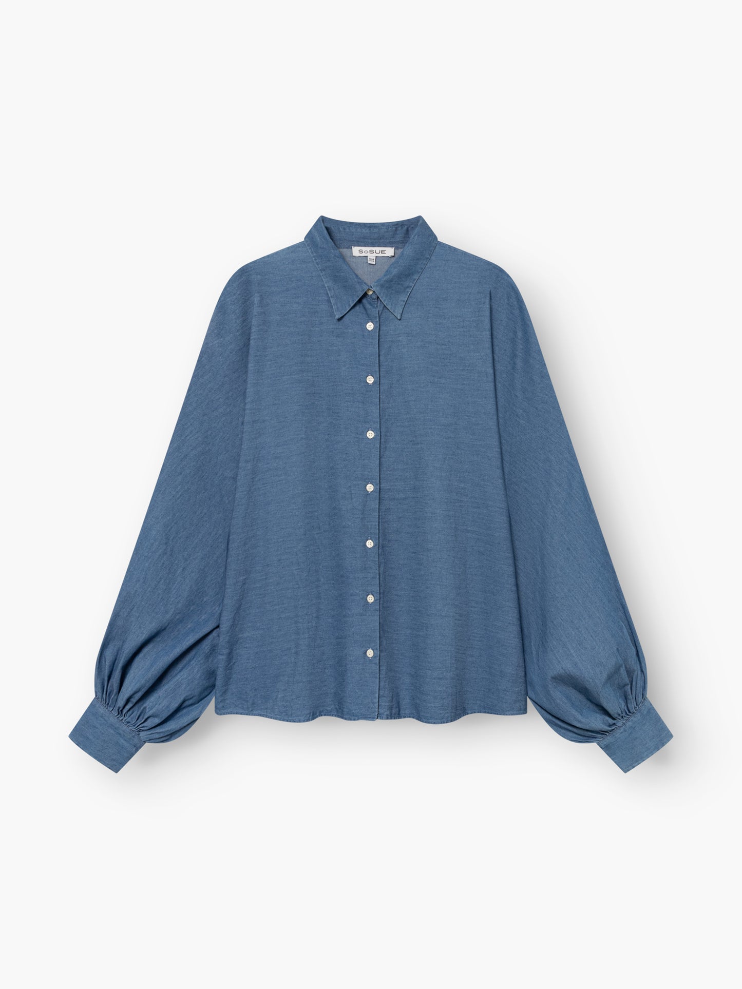Die Bluse „Antonia Denim“ ist eine locker sitzende blaue Jeansbluse mit langen Ärmeln, einem Kragen, weißen Knöpfen auf der Vorderseite und breiten, gerafften Ärmelbündchen, abgebildet vor weißem Hintergrund.