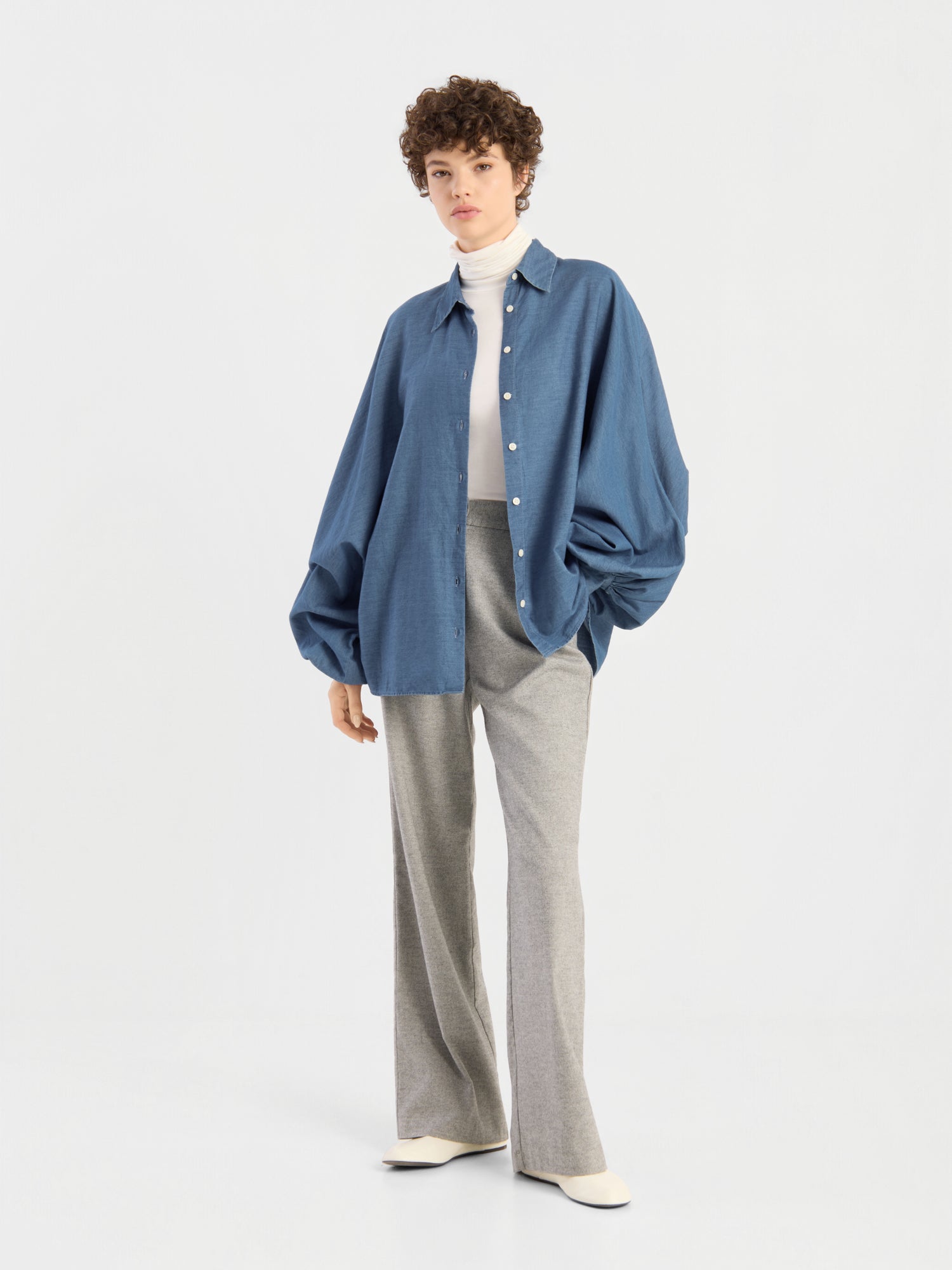 Eine Person steht vor einem schlichten Hintergrund und trägt eine übergroße Bluse von Antonia Denim über einem weißen Rollkragenpullover, dazu eine graue Hose mit weitem Bein und weiße Schuhe. Kurzes, lockiges Haar und eine entspannte Haltung runden den Look ab.