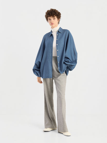 Eine Person steht vor einem schlichten Hintergrund und trägt eine übergroße Bluse von Antonia Denim über einem weißen Rollkragenpullover, dazu eine graue Hose mit weitem Bein und weiße Schuhe. Kurzes, lockiges Haar und eine entspannte Haltung runden den Look ab.