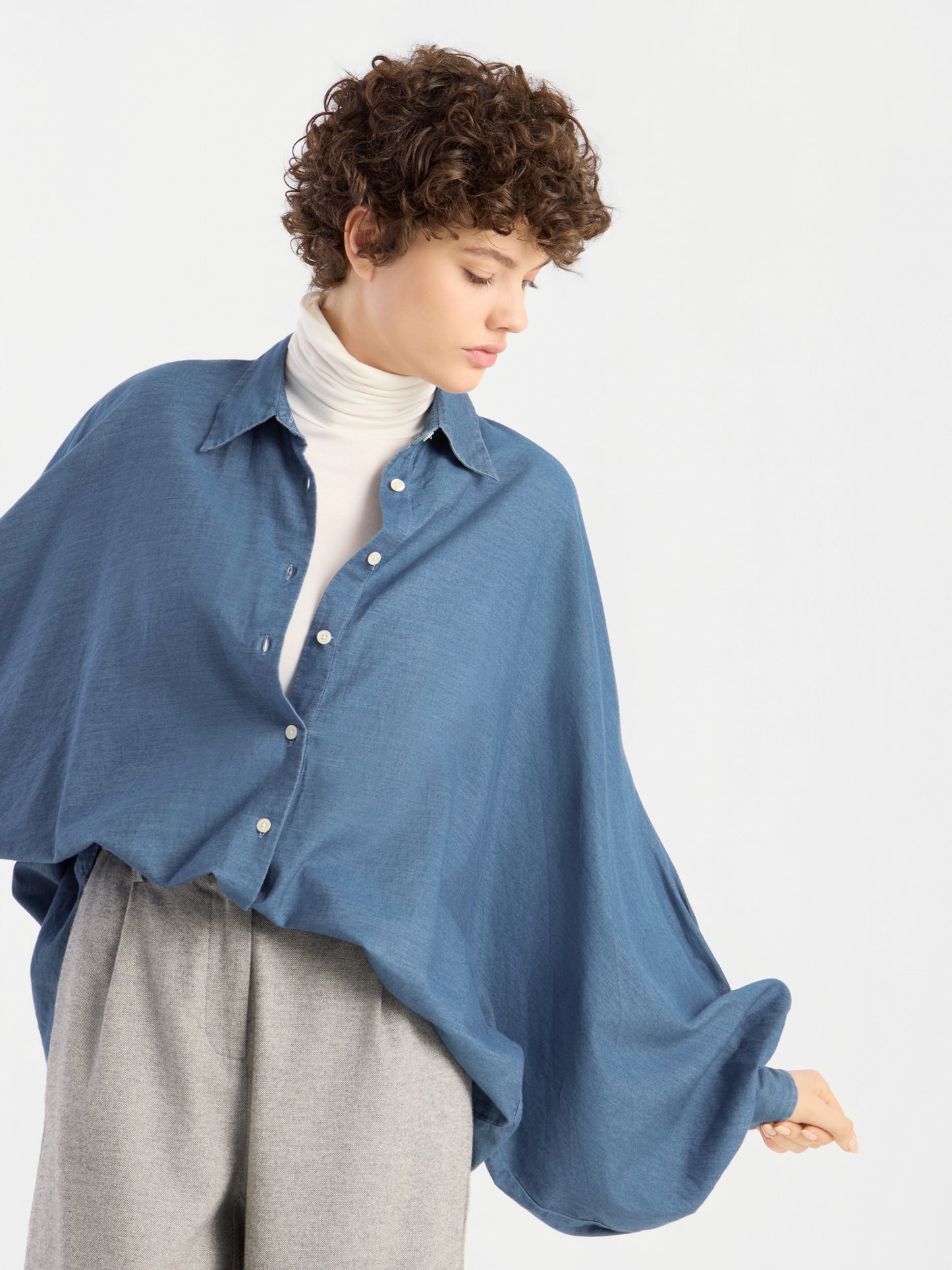 Eine Person mit kurzen Locken trägt eine Oversize-Bluse von „Bluse Antonia Denim“ über einem weißen Rollkragenpullover und einer grauen Hose; sie blickt nach unten und hält den Ärmel der Bluse vor einem schlichten, hellen Hintergrund hoch.