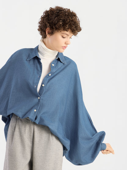 Eine Person mit kurzen Locken trägt eine Oversize-Bluse von „Bluse Antonia Denim“ über einem weißen Rollkragenpullover und einer grauen Hose; sie blickt nach unten und hält den Ärmel der Bluse vor einem schlichten, hellen Hintergrund hoch.