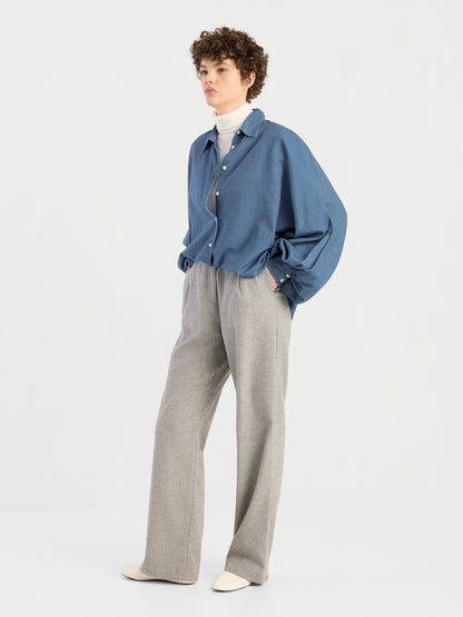 Eine Person mit kurzen Locken trägt die übergroße Bluse „Antonia Denim“ in Blau über einem weißen Rollkragenpullover, einer lockeren grauen Hose und weißen Schuhen und steht mit den Händen in den Taschen vor einem schlichten hellen Hintergrund.