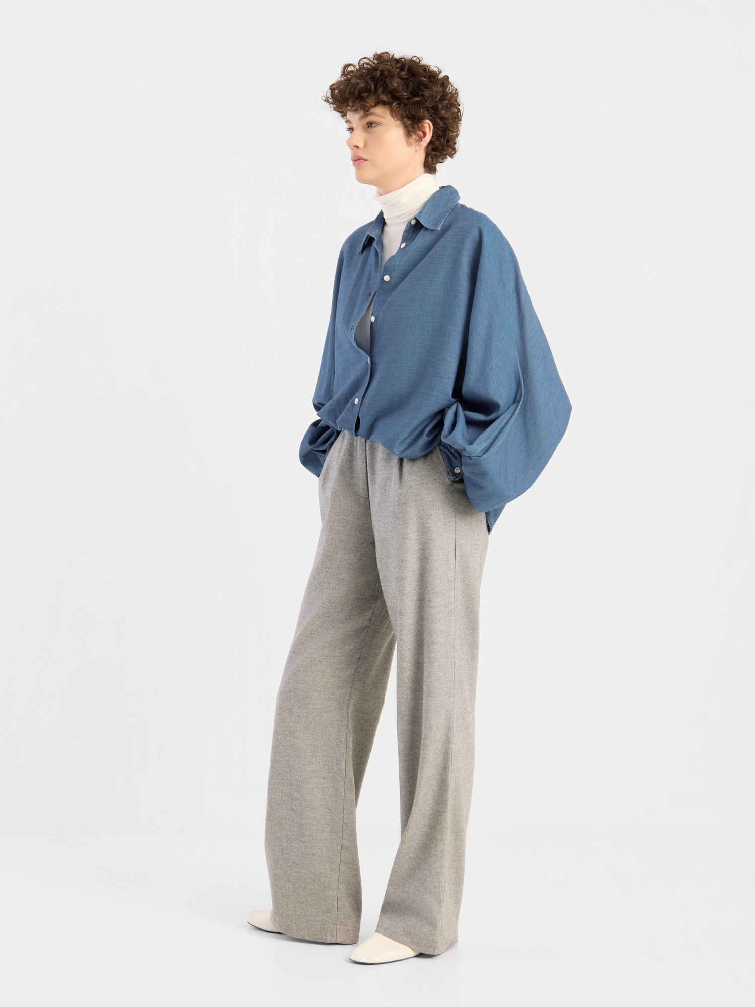 Vor einem schlichten, hellen Hintergrund steht eine Person mit kurzen Locken, die die Hände in den Taschen hat und eine übergroße Bluse von Antonia Denim über einem weißen Rollkragenpullover, eine graue Hose mit weitem Bein und weiße Schuhe trägt.