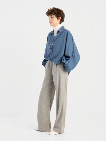 Vor einem schlichten, hellen Hintergrund steht eine Person mit kurzen Locken, die die Hände in den Taschen hat und eine übergroße Bluse von Antonia Denim über einem weißen Rollkragenpullover, eine graue Hose mit weitem Bein und weiße Schuhe trägt.