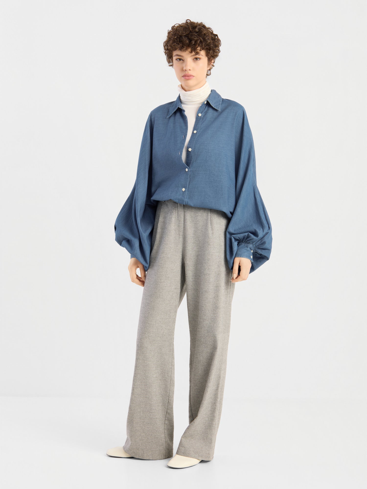 Eine Person mit kurzen Locken trägt die Bluse „Antonia Denim“ in Oversize-Passform über einem weißen Rollkragenpullover, kombiniert mit einer grauen Hose mit weitem Bein und beigen Schuhen, vor einem schlichten weißen Hintergrund. Der Look unterstreicht den Tragekomfort von 100 % Baumwolle.