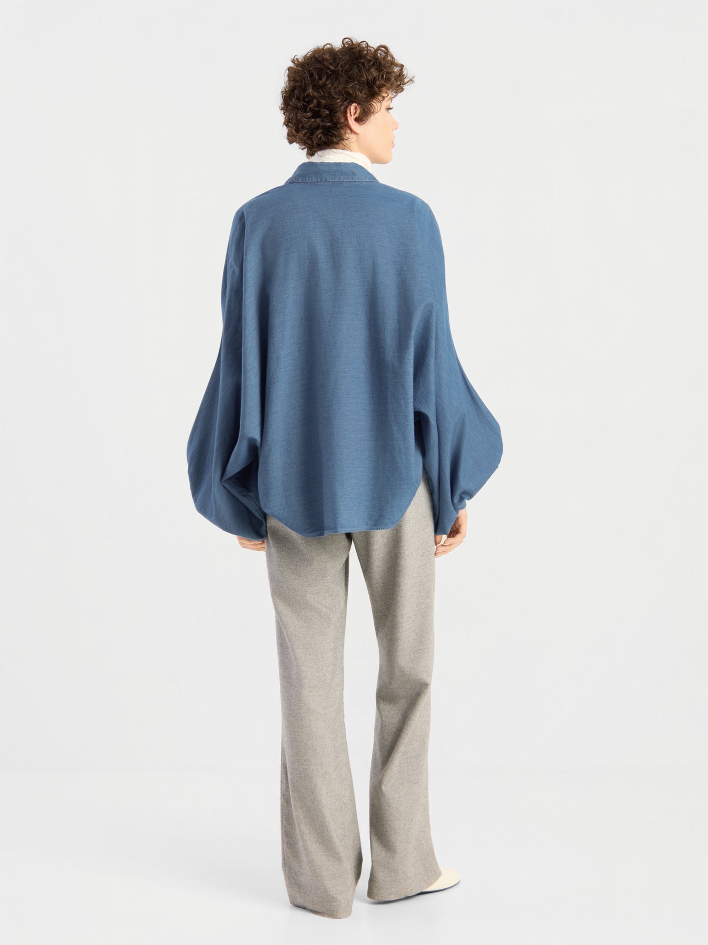 Eine Person mit kurzen Locken blickt in eine andere Richtung und trägt eine Bluse von Antonia Denim in Oversize-Passform mit weiten Ärmeln sowie eine hellgraue Hose mit weitem Bein, vor einem schlichten weißen Hintergrund.