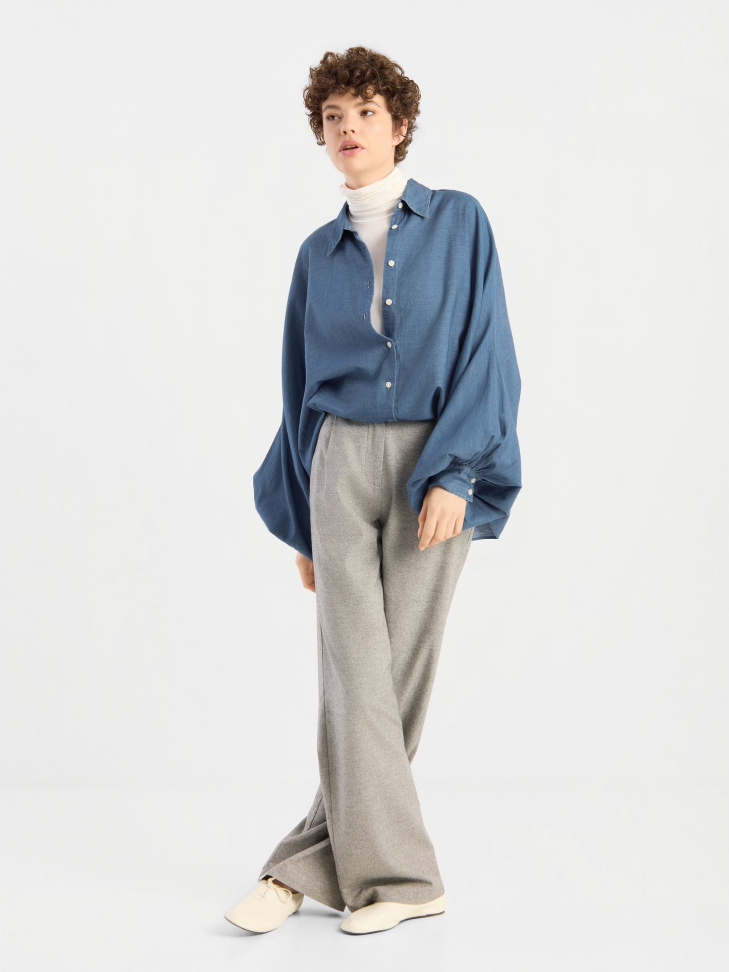 Eine Person mit kurzen Locken trägt eine übergroße Bluse von Antonia Denim über einem weißen Rollkragenpullover und einer grauen Hose mit weitem Bein, dazu cremefarbene Schuhe, und steht vor einem schlichten, hellen Hintergrund.