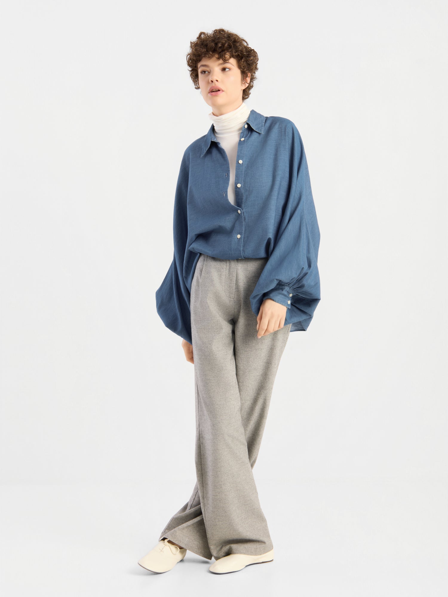 Eine Person mit kurzen Locken trägt eine übergroße Bluse von Antonia Denim über einem weißen Rollkragenpullover und einer grauen Hose mit weitem Bein, dazu cremefarbene Schuhe, und steht vor einem schlichten, hellen Hintergrund.