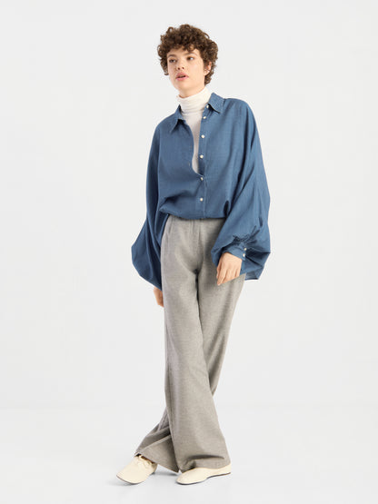 Eine Person mit kurzen Locken trägt eine übergroße Bluse von Antonia Denim über einem weißen Rollkragenpullover und einer grauen Hose mit weitem Bein, dazu cremefarbene Schuhe, und steht vor einem schlichten, hellen Hintergrund.