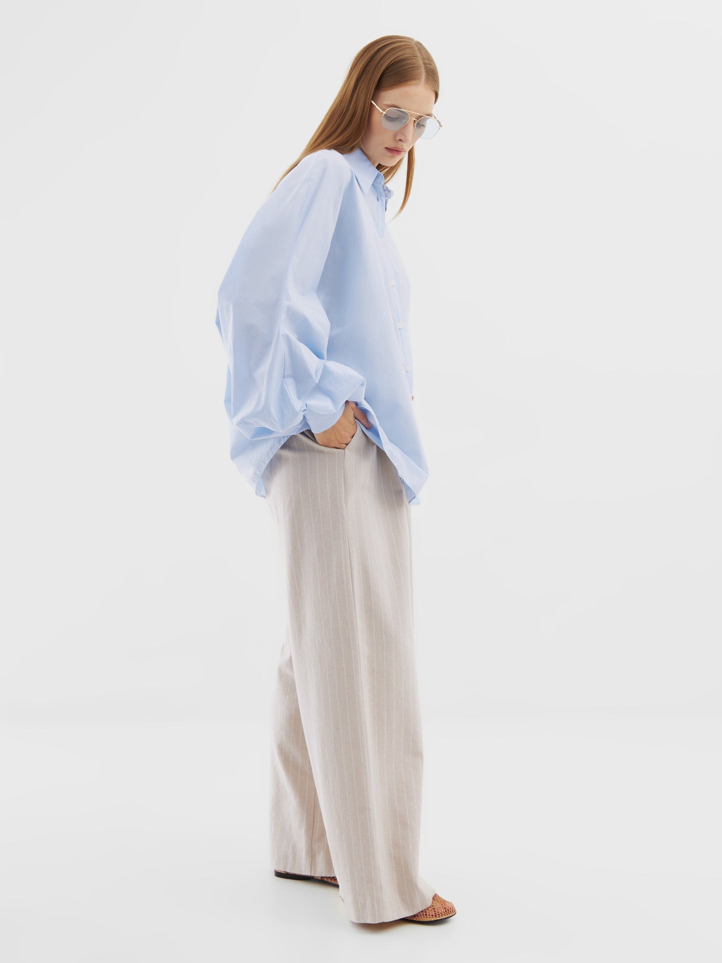 Eine Frau, die die Bluse „Antonia Hellblau“ aus Baumwolle, eine beige Hose mit weitem Bein, eine Sonnenbrille und Sandalen trägt, steht mit den Händen in den Taschen vor einem schlichten weißen Hintergrund.